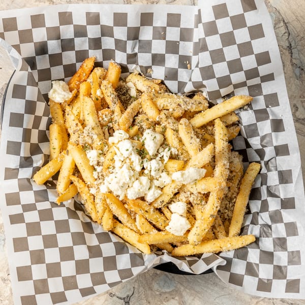 A basket of Krazy Greek fries, topped with crumbled feta cheese and herbs, on checkered paper.