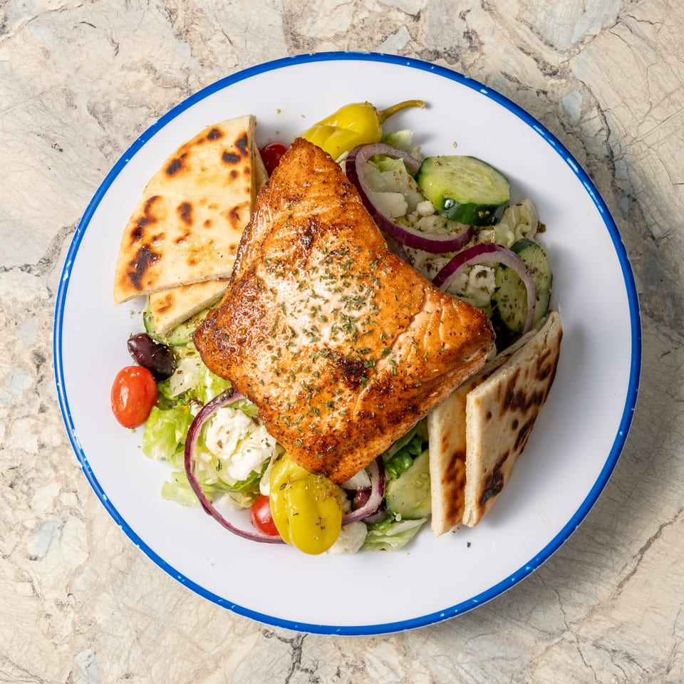 Krazy Greek salmon entree with Greek salad, pita bread, and a pepperoncini on a white plate.